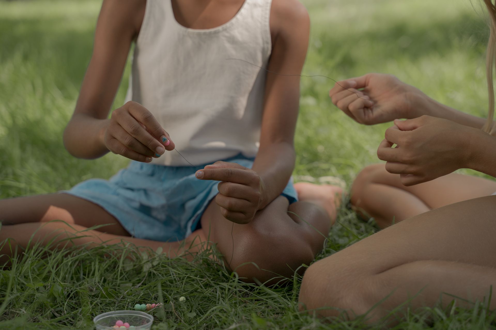 Two children sit in a park making bead necklaces, enjoying a summer day.