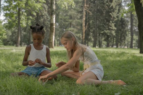 Free Two girls sitting on grass creating bead necklaces in a sunny park. Stock Photo