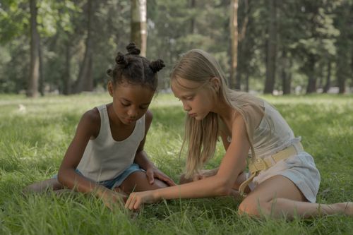 Free Two girls creating bead necklaces while sitting on grass in a sunny park. Stock Photo