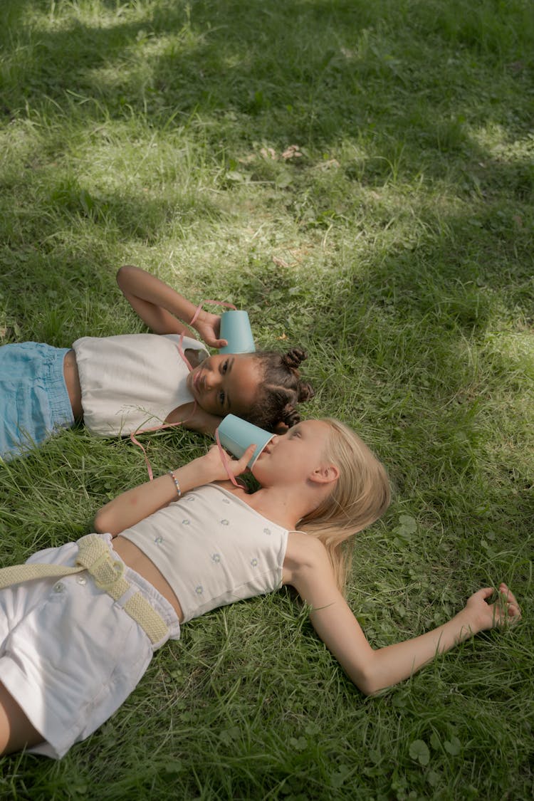 Two Girls Laying On Lawn And Playing Paper Cups Connected With String