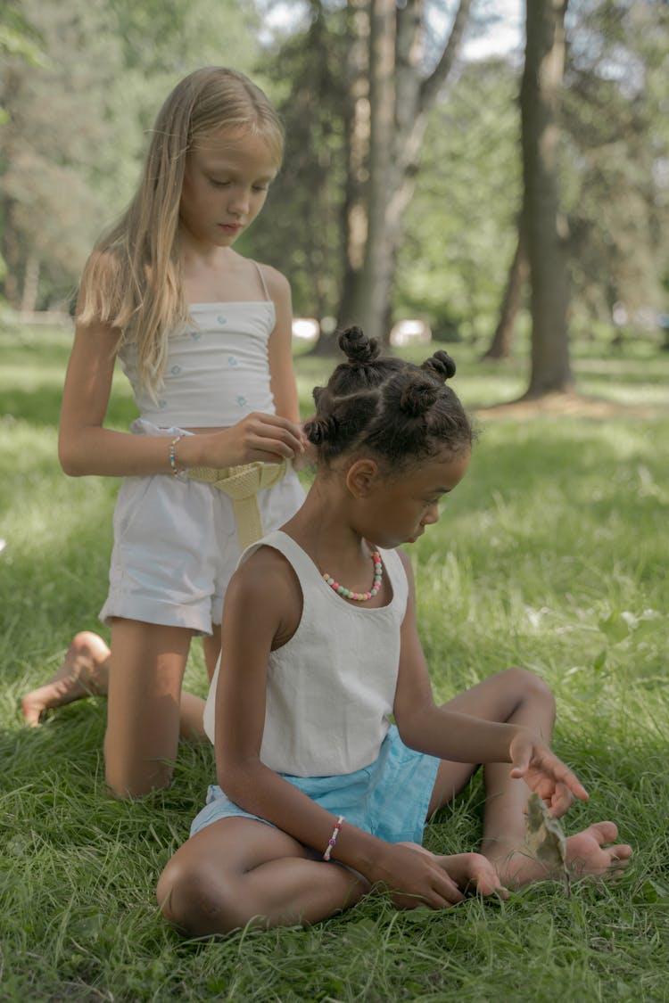 Blond Teenage Girl Kneeling And Doing Hair Of Girl Sitting On Grass In Park