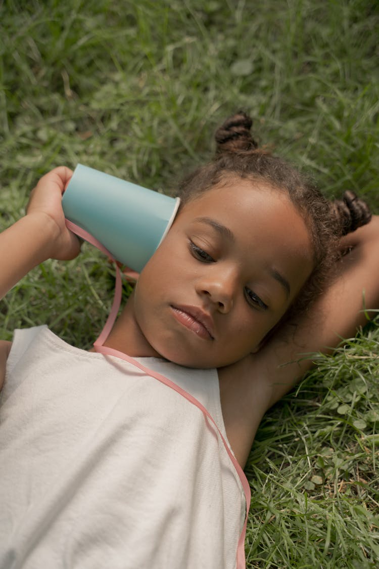 Teenage Girl Laying On Grass And Holding Blue Paper Cup At Ear