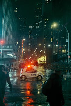 Moody NYC street scene featuring NYPD car and pedestrians under umbrellas with neon lights reflecting on wet pavement.