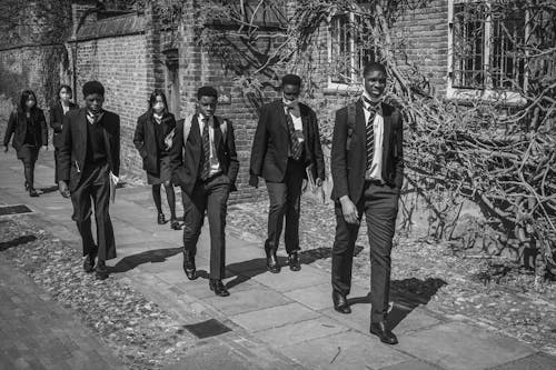 Grayscale Photo of a Group of Men in Black Suit and Women Walking on Sidewalk