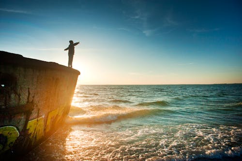 Free Silhouette of Person on Cliff Beside Body of Water during Golden Hour Stock Photo