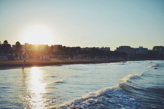 A person running along a sunlit beach at sunset with gentle waves and buildings in the background.