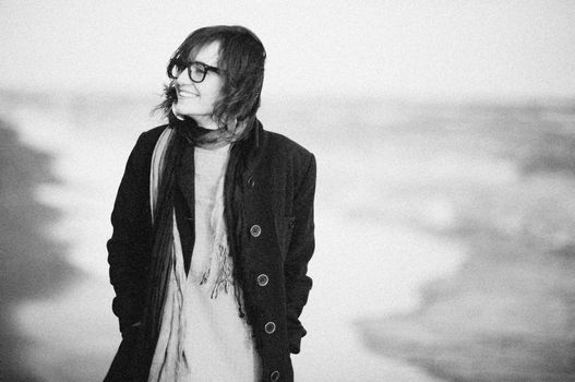Charming black and white image of a smiling woman in a coat on a beach. Timeless and serene vibe.