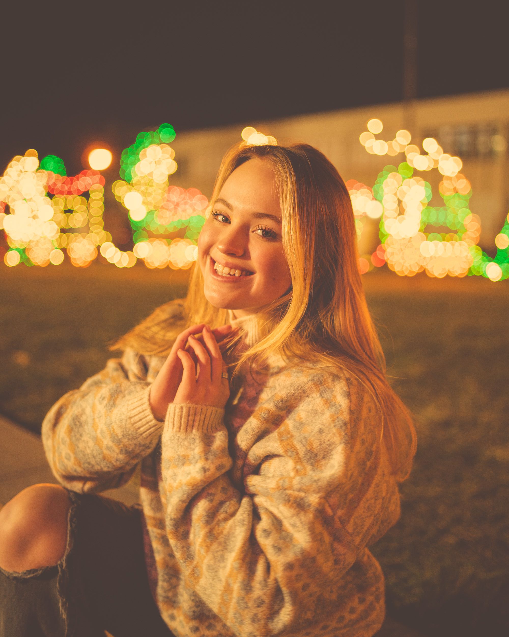 A woman enjoying the festive glow of holiday lights outdoors at night, wearing a cozy sweater and smiling brightly.