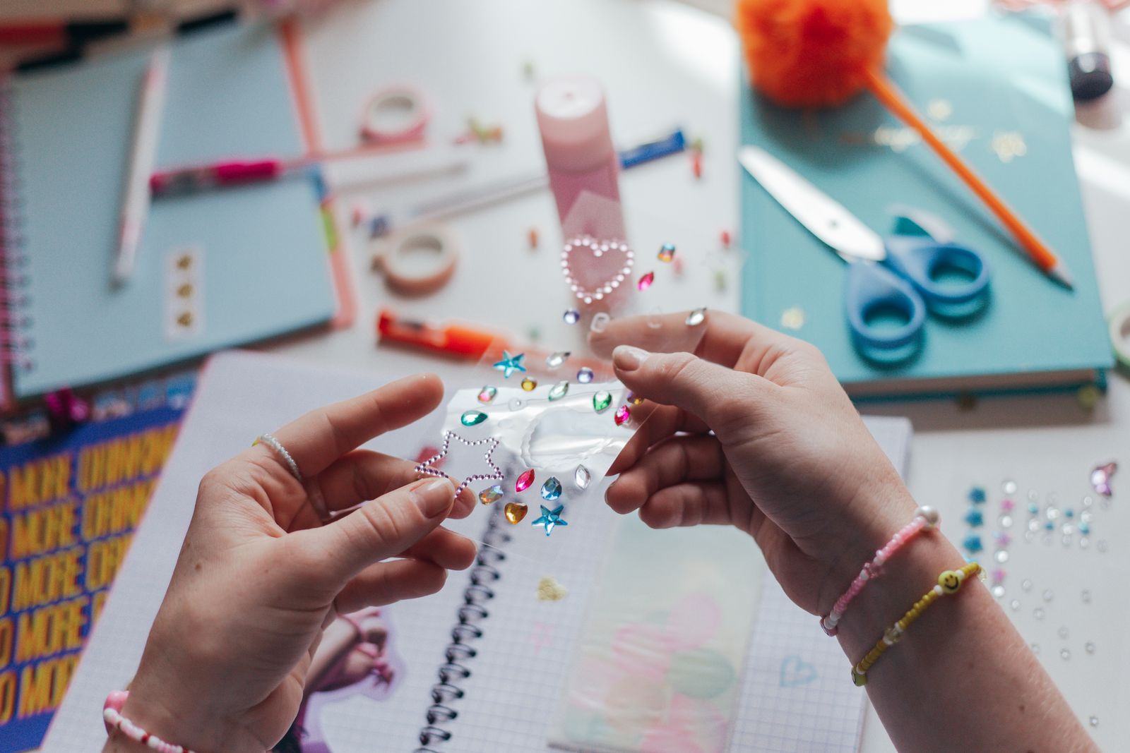 Hands peeling a sheet of colourful rhinestone stickers to decorate a notebook on a desk scattered with craft supplies