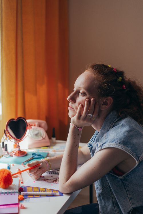 Free Caucasian woman in denim vest pondering at desk with art supplies, reflecting creativity and contemplation. Stock Photo