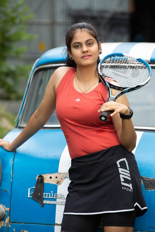 A Woman in Red Tank Top Holding a Tennis Racket while Leaning on a Blue Car
