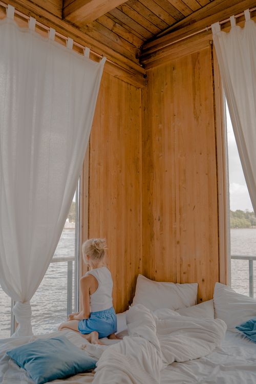 Free A child sitting on a bed, looking through a window in a warm wooden bedroom setting. Stock Photo