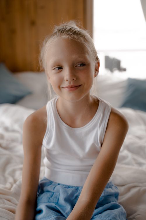 Free A cheerful young girl with blond hair smiling while sitting on a bed in a bright bedroom. Stock Photo