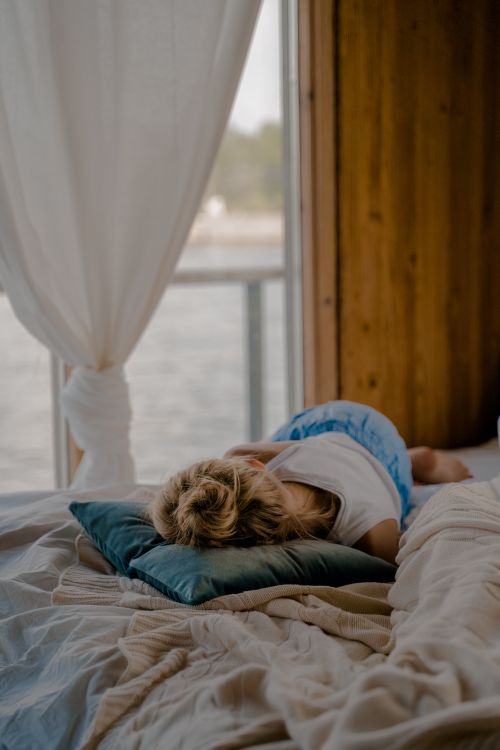 Free A young girl peacefully rests on a bed in a wooden cabin, enjoying a serene lake view. Stock Photo