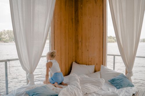 Free A child sits on a bed looking out of a window, enjoying a serene view. Relaxing indoor setting. Stock Photo