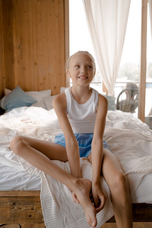 Free A smiling girl with blond hair sits cheerfully on a bed in a cozy, bright bedroom. Stock Photo