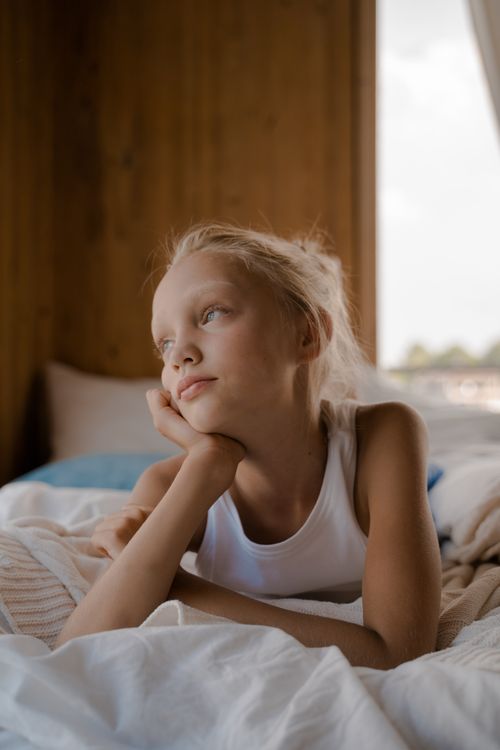 Free A young girl with blond hair lying on a bed, gazing thoughtfully. Stock Photo
