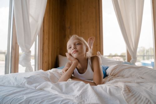 Free A child with blond hair lies relaxed on a bed inside a wooden room, enjoying a peaceful moment. Stock Photo