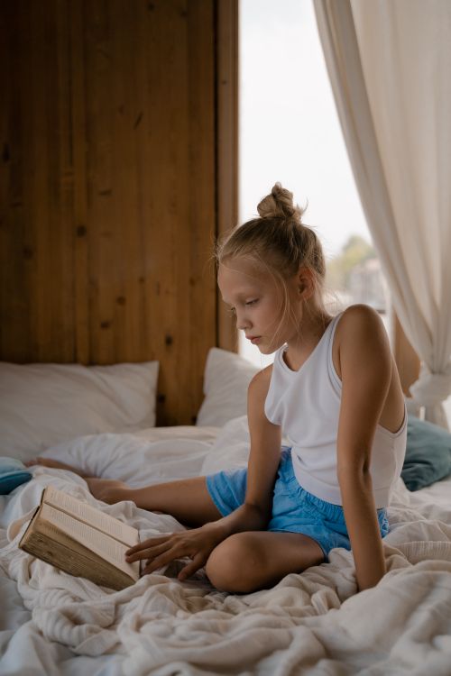 Free A young girl with blond hair sits on a bed, engrossed in reading, creating a serene and cozy atmosphere. Stock Photo