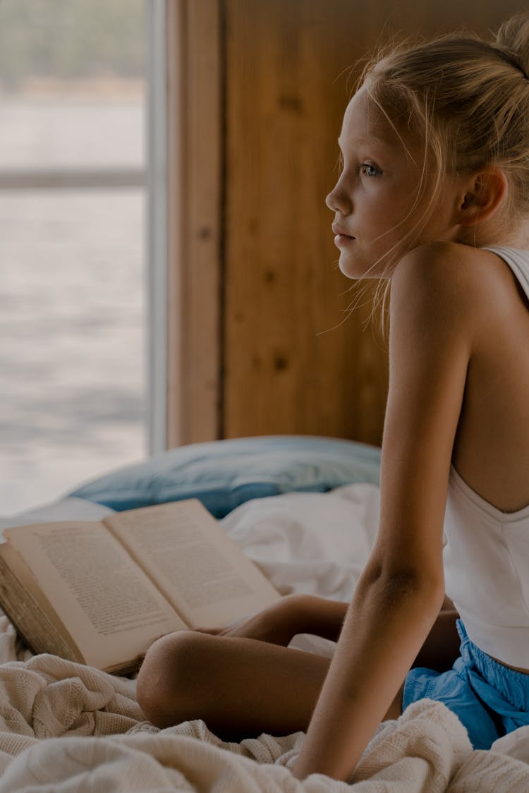 Sad Child Sitting On Bed With Book