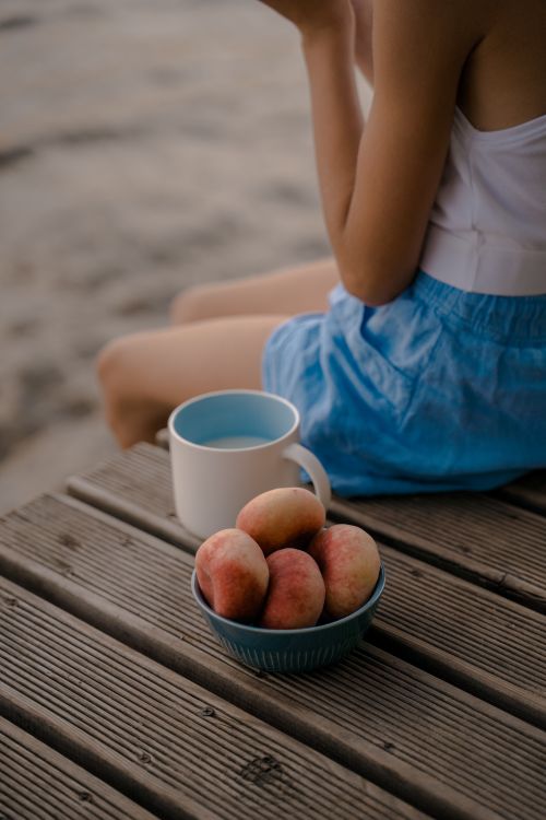 Free A young woman enjoys a serene morning by the pier with a bowl of peaches and a mug of milk. Stock Photo
