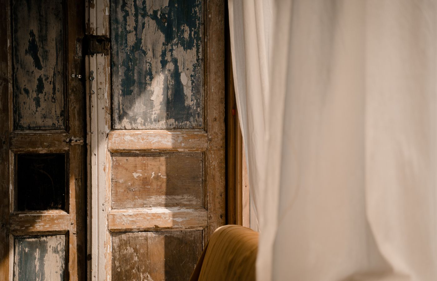 Sunlit rustic wooden door with aged texture partially covered by flowing curtains.