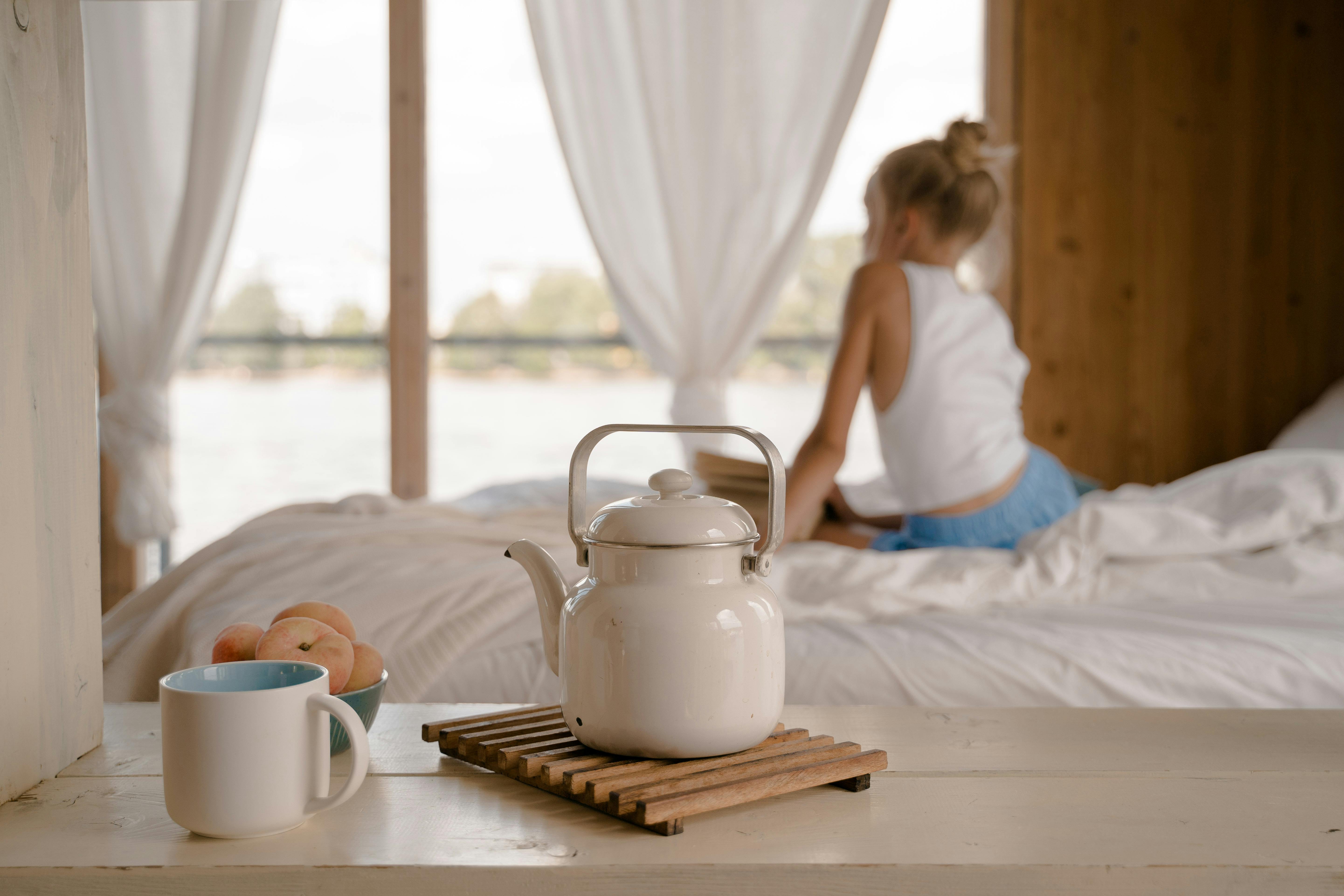 A relaxing morning scene with a teapot, mug, and fruit in a cozy bedroom setting.