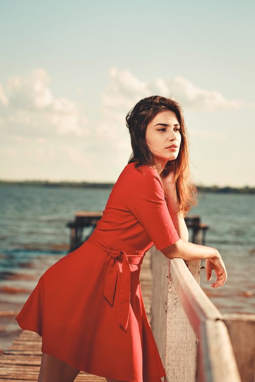 Free A woman in a red dress posing gracefully on a wooden dock by a serene lake during a bright day. Stock Photo