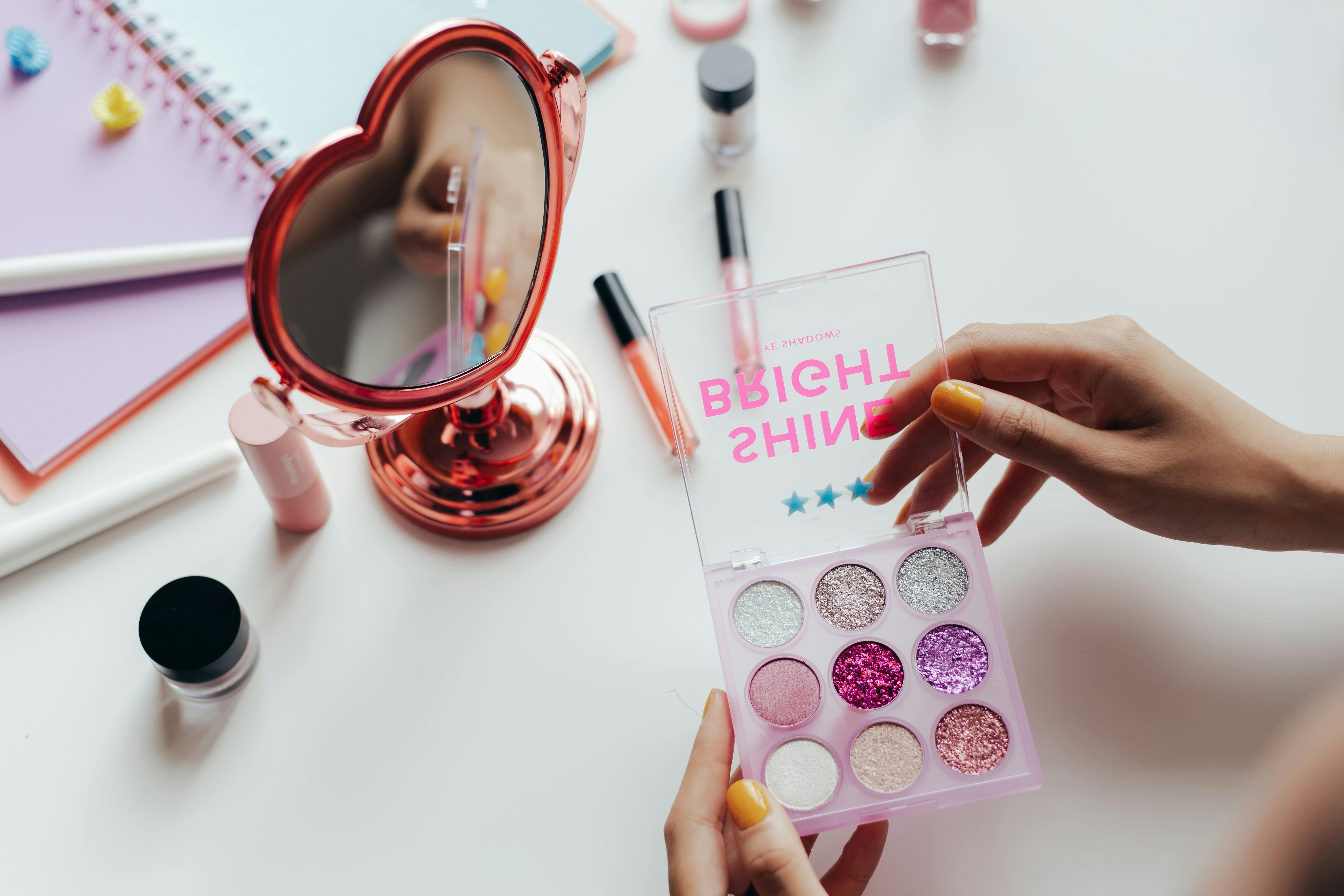 A vibrant eyeshadow palette with glittery colors held near a heart-shaped mirror.