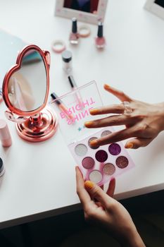 Close-up of a woman's hands using an eyeshadow palette with a heart-shaped mirror on a white table.