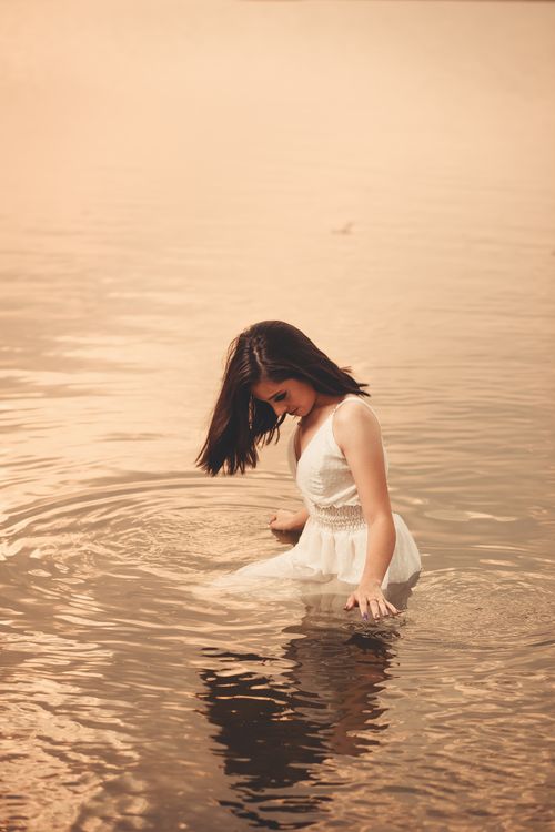 Free A woman in a white dress gracefully poses while standing in calm water, creating a peaceful and dreamy atmosphere. Stock Photo