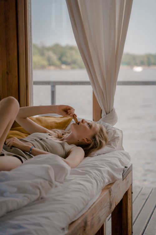 Free Woman enjoying a snack while lying on bed in a beach house by the water. Serene and peaceful atmosphere. Stock Photo