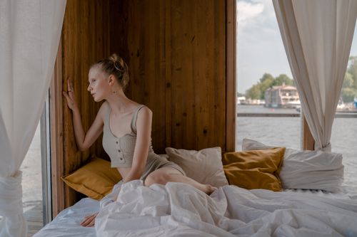 Free Woman sitting on bed and looking through window in a cozy wooden room. Stock Photo