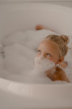 Cute child with blond hair having fun with bubbles in a bathtub, capturing a playful moment indoors.