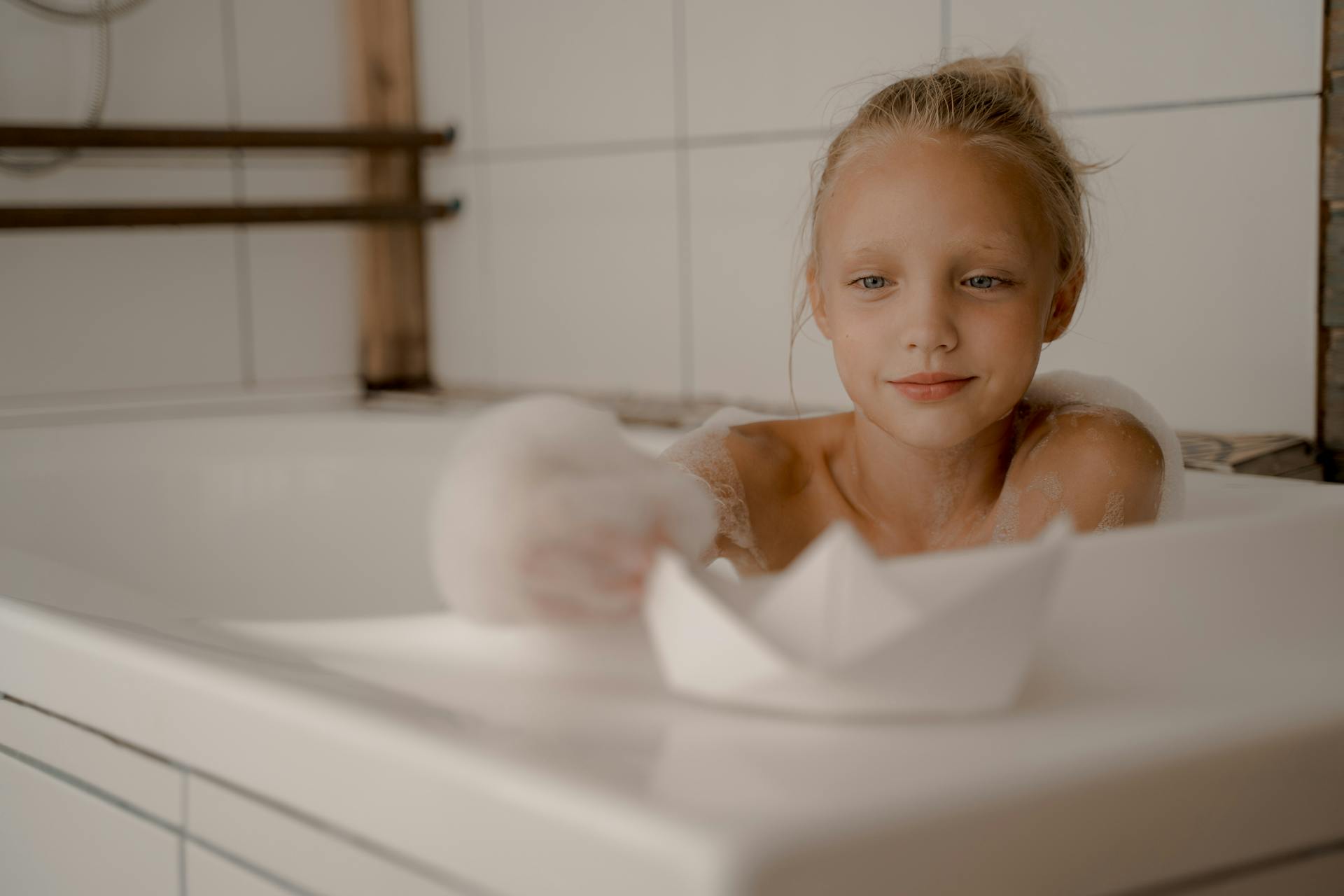 Blond girl having fun with a paper boat in a bathtub filled with bubbles.