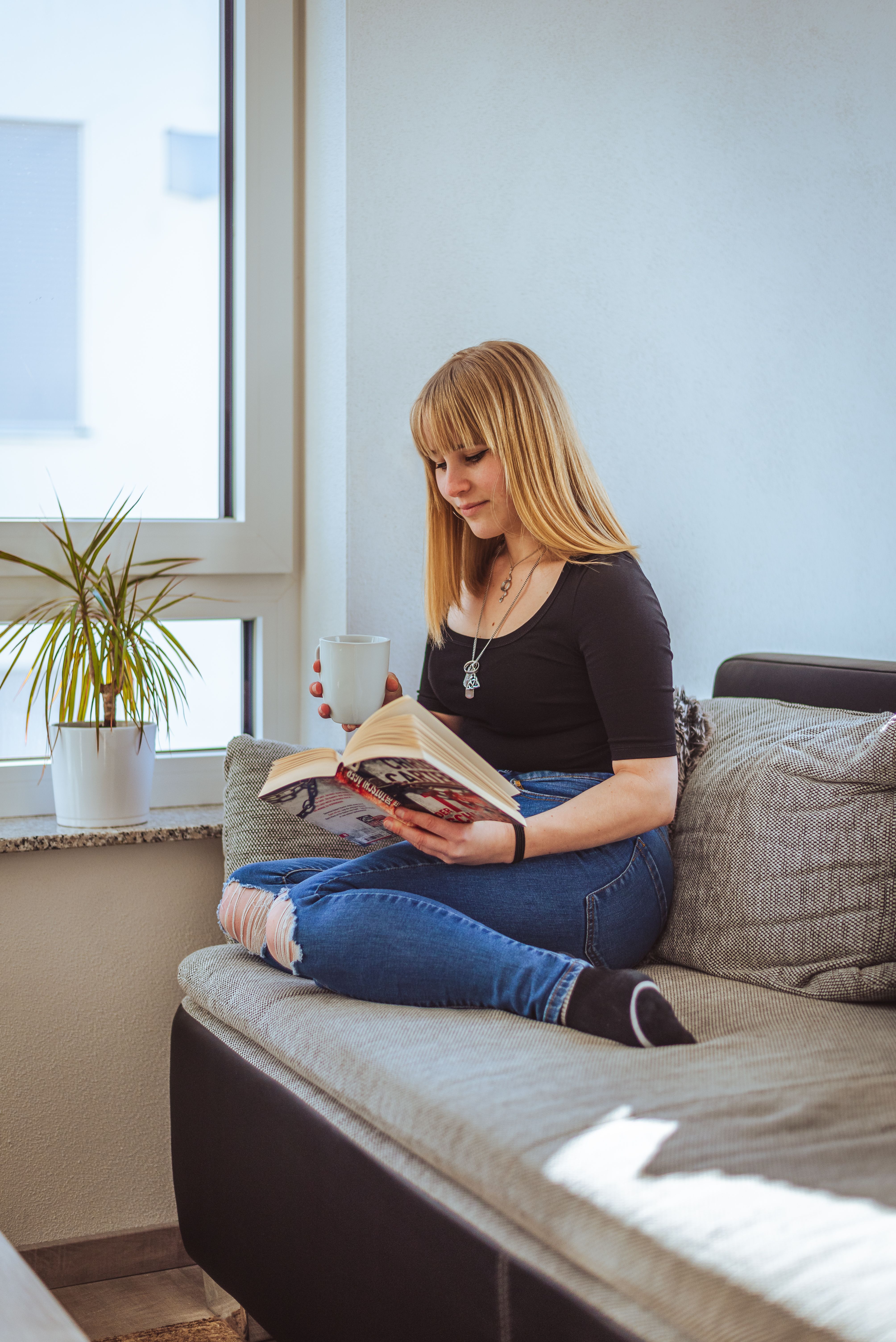 Relaxed young woman reading on a couch, sipping coffee in a sunny room.