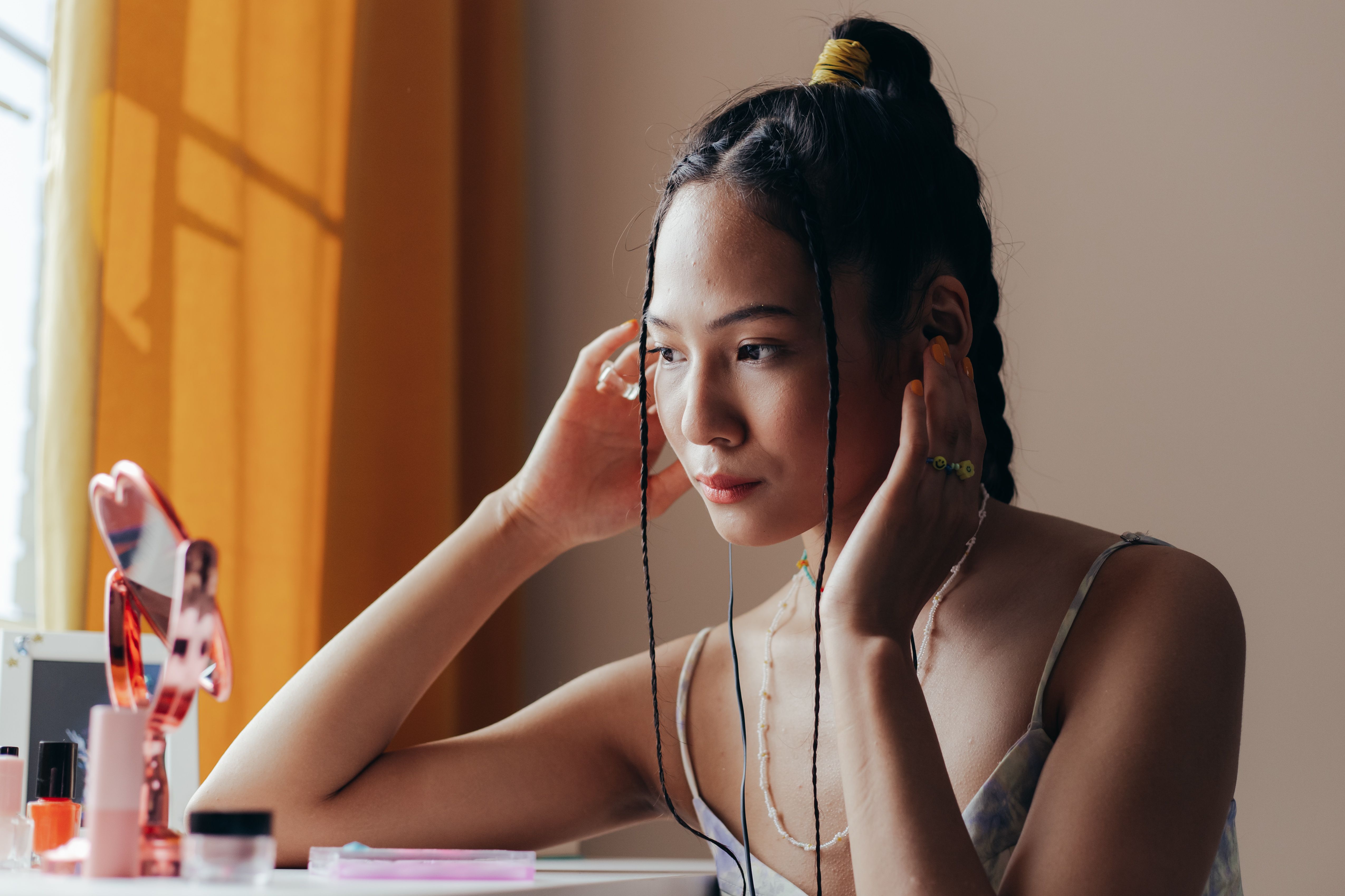 A woman with braided hair sitting by a mirror, applying earrings in a softly lit room.
