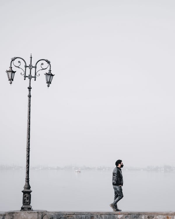 Photograph of a Man in a Black Jacket Walking Near a Black Street Lamp