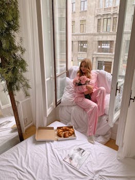 Woman in pink pajamas holding flowers while relaxing by an open window with croissants nearby in a light-filled room.