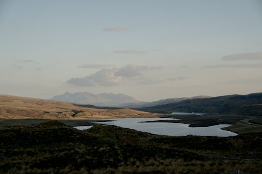 A tranquil view of a lake surrounded by hills and distant mountains during dusk.