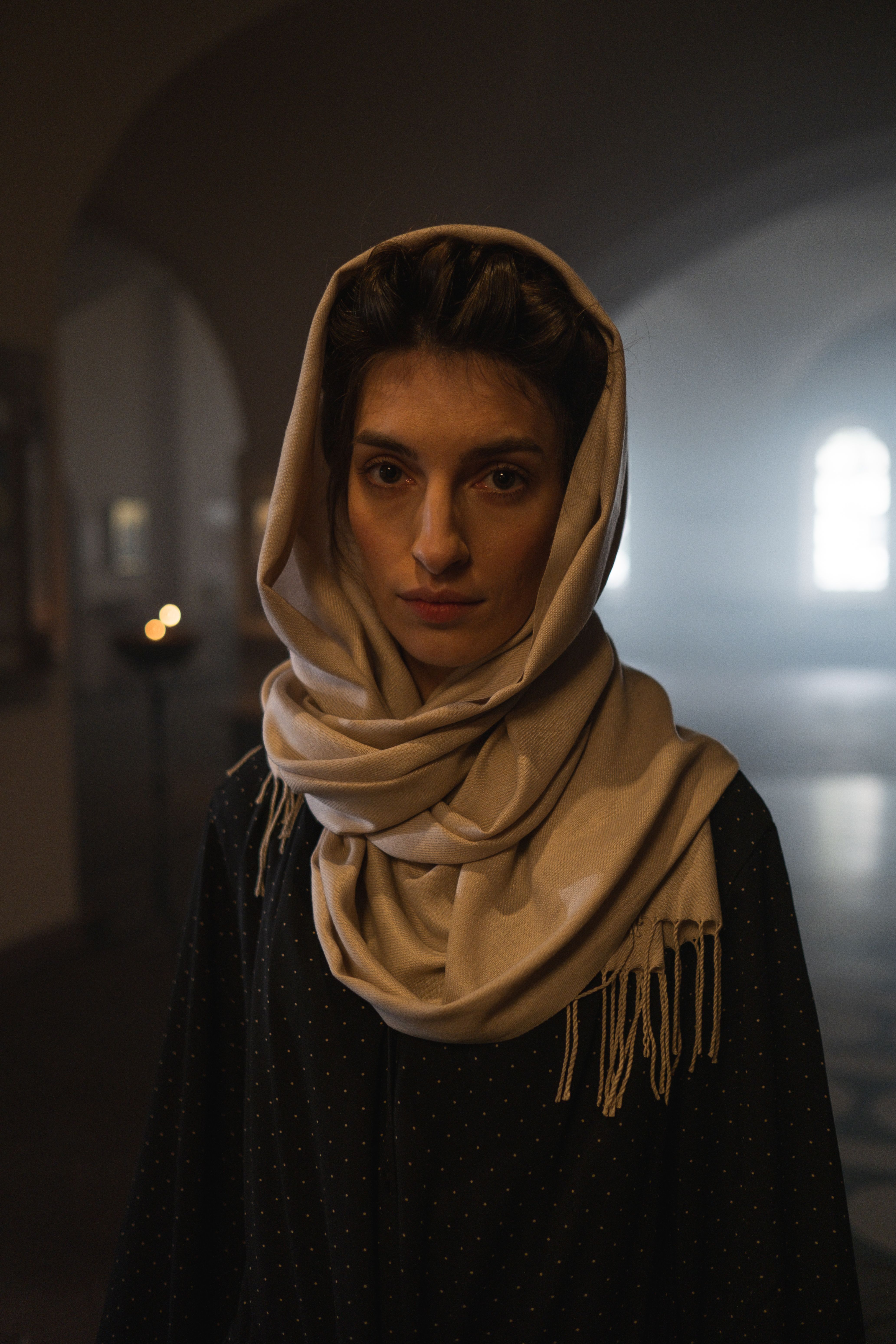 A contemplative woman in a headscarf stands inside a dimly lit church, enveloped in solemn ambience.
