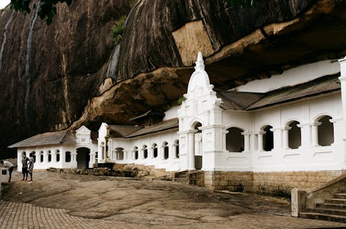 Free White architectural structure set against a rocky cliff with people exploring the site. Stock Photo