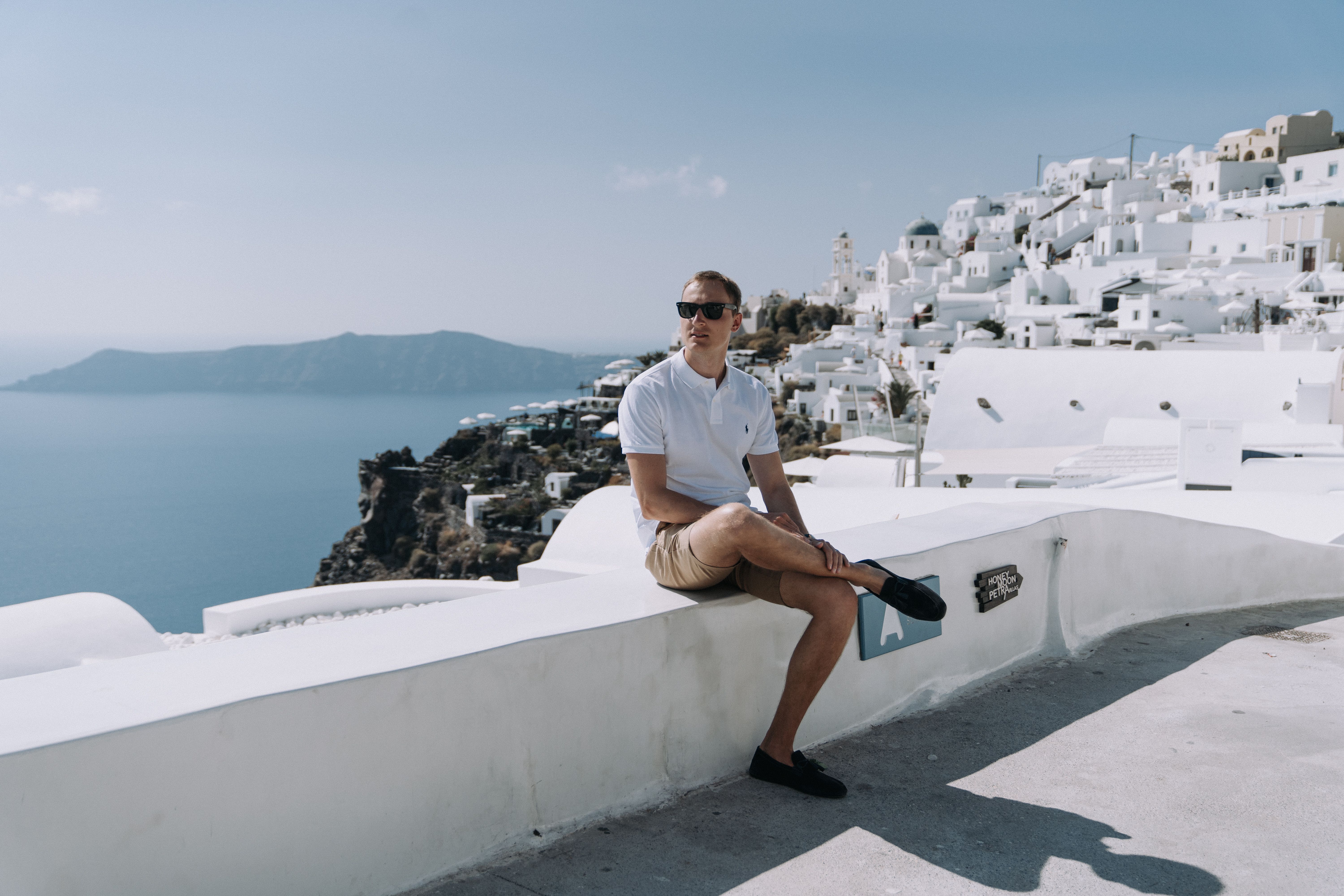 A man in sunglasses and white polo enjoys the scenic view of Santorini's coastline from a concrete bench.