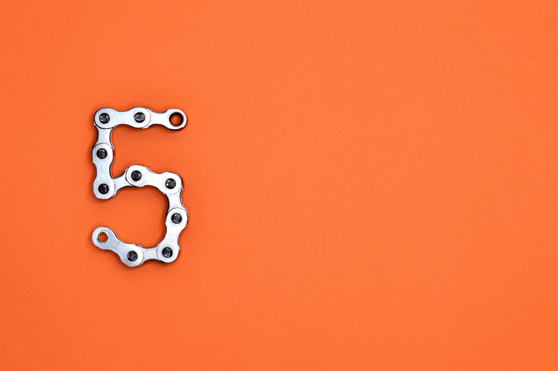 Creative arrangement of a chrome bicycle chain forming the number 5 on a vibrant orange background.