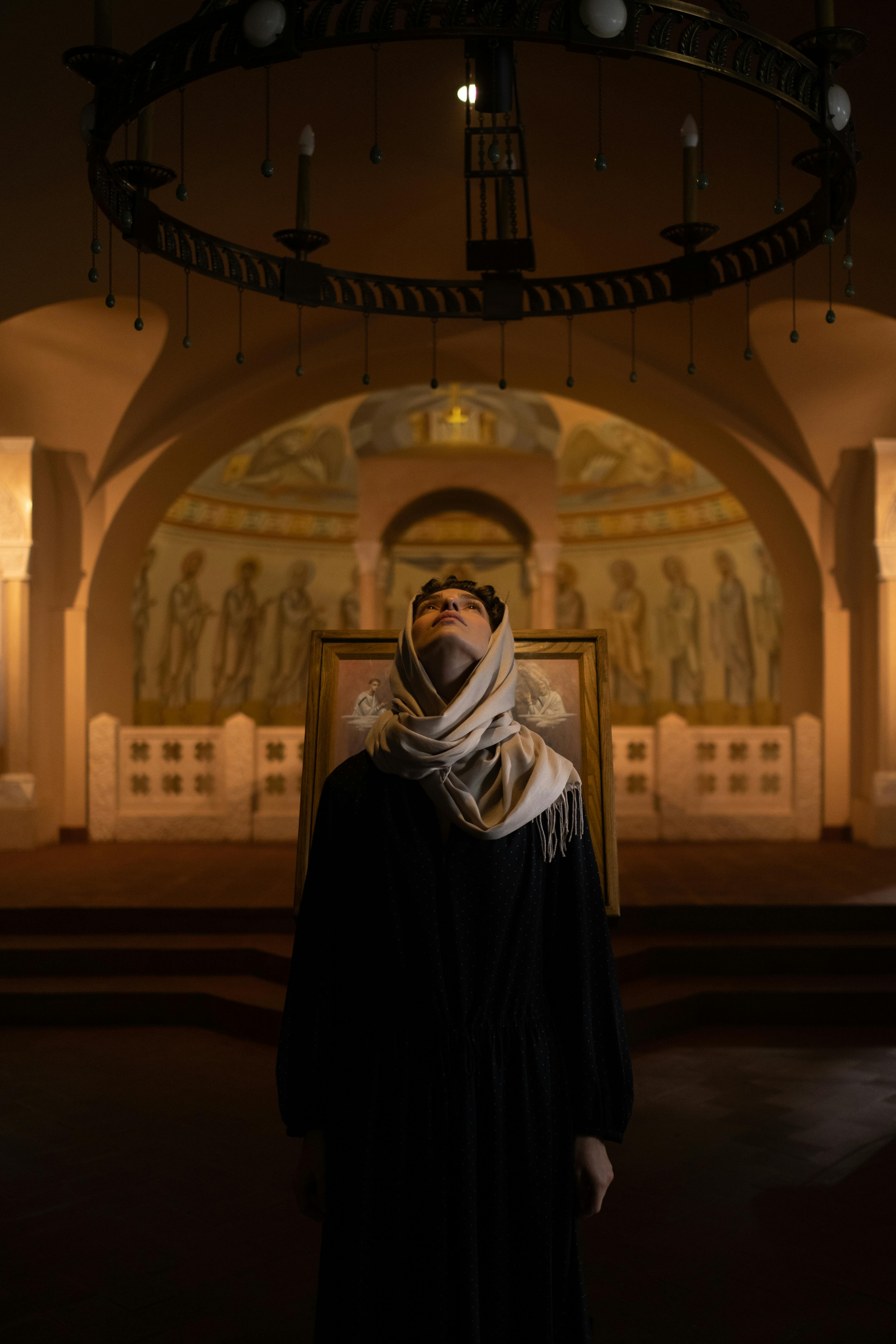 Woman with headscarf contemplating beneath ornate church chandelier.
