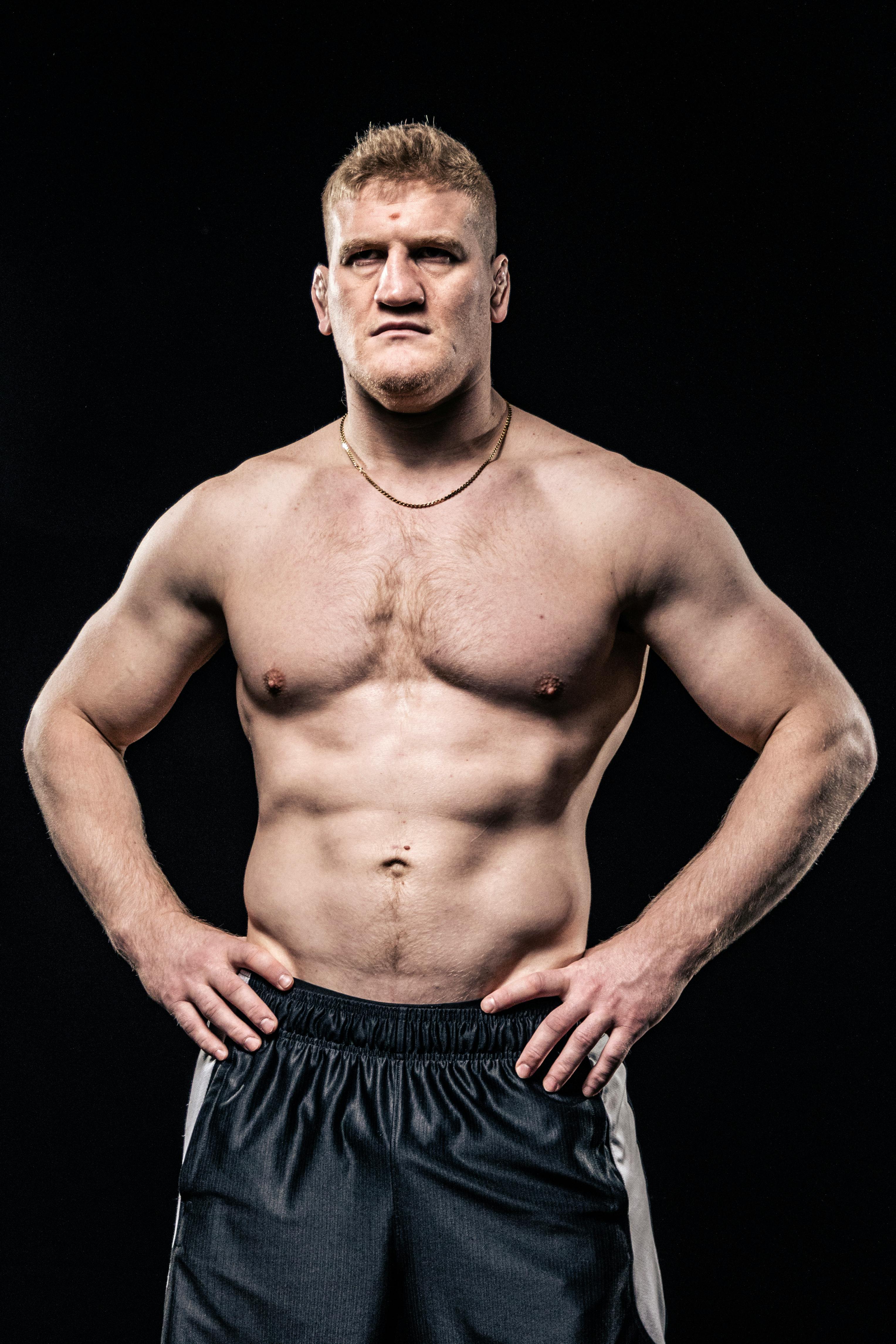 Shirtless bodybuilder showcasing muscular physique with intense expression on dark background.