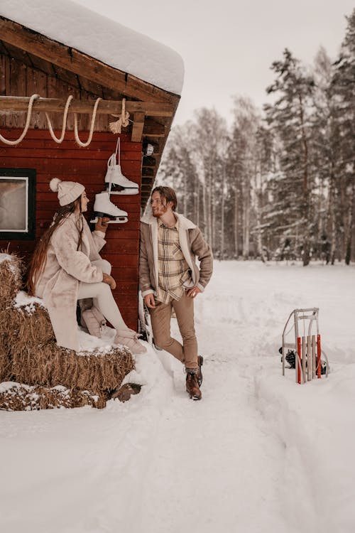 A Woman Sitting Near the Wooden House Near Her Partner Standing on a Snow Covered Ground