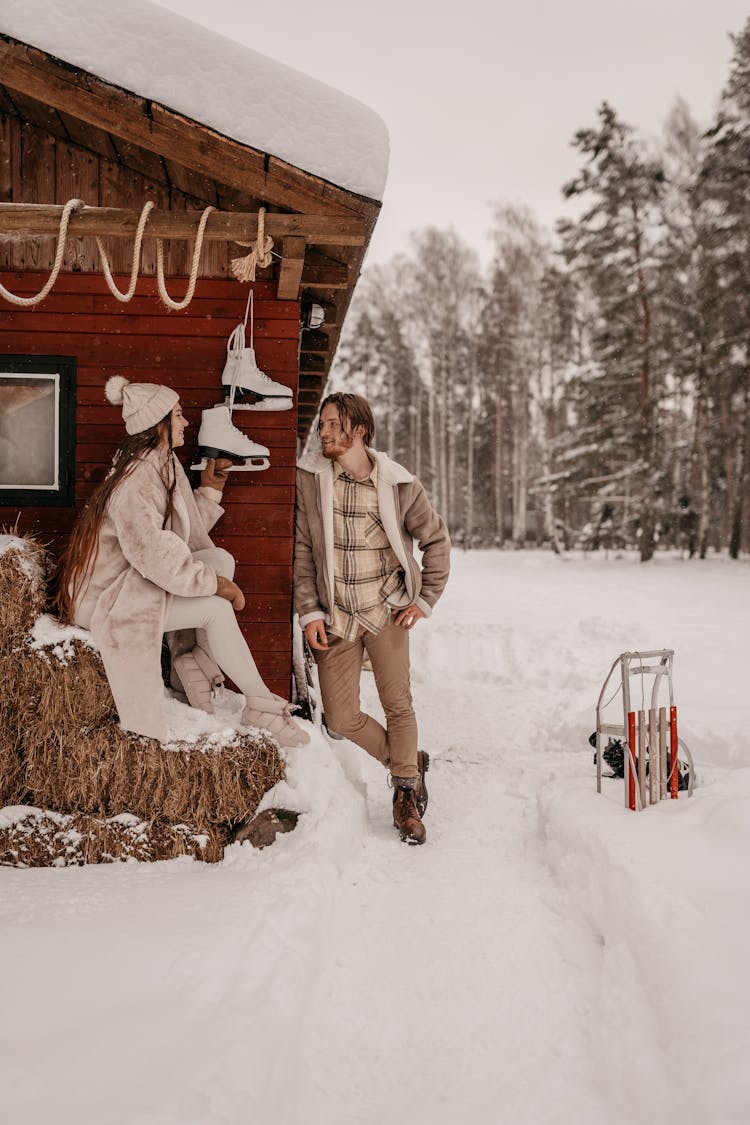 A Woman Sitting Near The Wooden House Near Her Partner Standing On A Snow Covered Ground