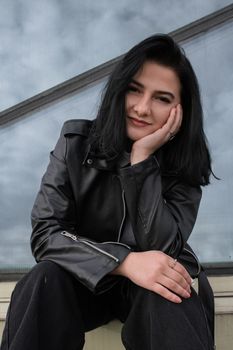 Confident woman in a black leather jacket sitting outdoors with a thoughtful smile.
