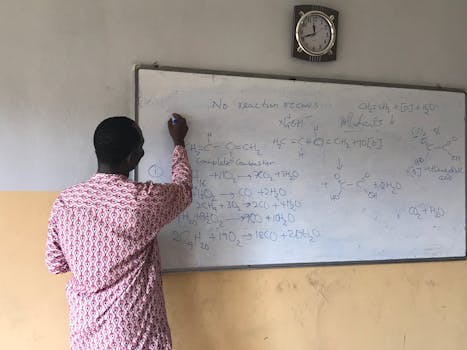 Male teacher writing chemical equations on a classroom whiteboard.